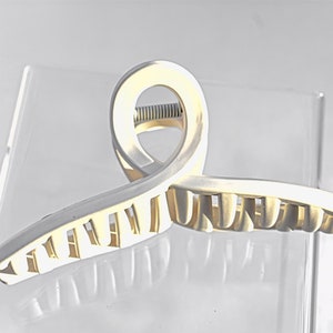 Puede incluir: Una pinza para el pelo de plástico blanco con un mecanismo de resorte. La pinza tiene una forma única con un bucle abierto curvado en la parte superior y una fila de dientes en la parte inferior.