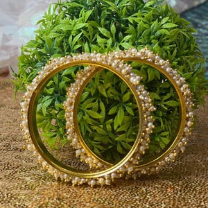 May include: Two gold bangles with white pearl accents. The bangles are stacked on top of each other.