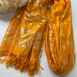 Puede incluir: Una bufanda amarilla con un patr&oacute;n geom&eacute;trico dorado y blanco y flecos.