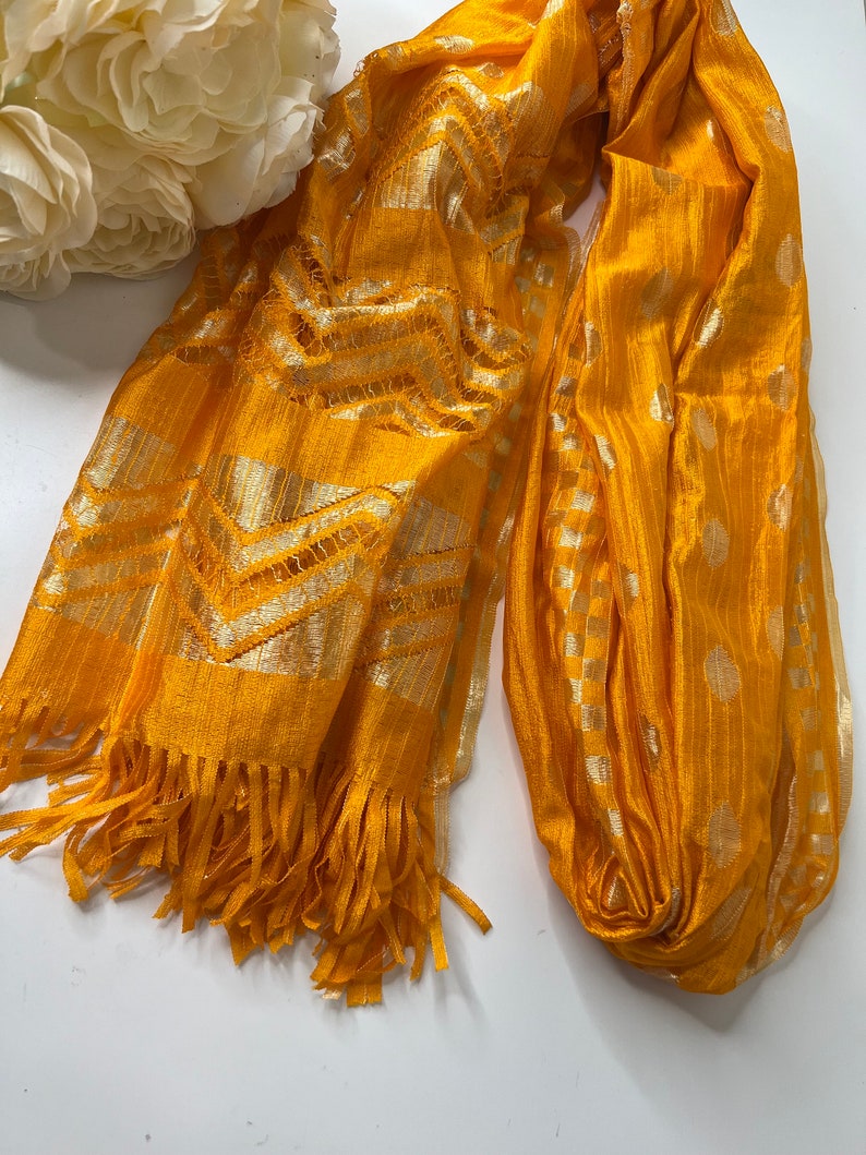 Puede incluir: Una bufanda amarilla con un patr&oacute;n geom&eacute;trico dorado y blanco y flecos.