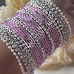 May include: A stack of lavender bangles with a textured design. The bangles are accented with rows of white pearls and silver-toned embellishments. The jewelry is displayed against a soft, blurred background.