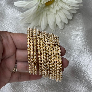 May include: A set of gold bangles with white beads. The bangles are stacked on top of each other and are being held in a hand.
