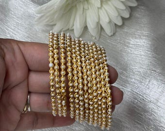 Pode incluir: Um conjunto de pulseiras douradas com contas brancas. As pulseiras estão empilhadas umas sobre as outras e estão sendo seguradas em uma mão.
