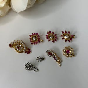 May include: A set of seven gold and silver nose rings with pink and white gemstones. The nose rings are in various shapes and sizes, including a round shape, a square shape, and a flower shape.