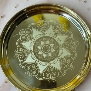 May include: A gold-colored, round decorative tray with an intricate floral design etched in a lighter gold hue. The tray has a slightly raised edge and a polished finish, suitable for serving or display.