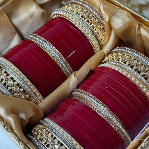 May include: A set of red bangles with gold and silver embellishments, presented in a gold box. The bangles feature intricate designs with pearls and rhinestones, creating a luxurious look. The box is lined with gold fabric.