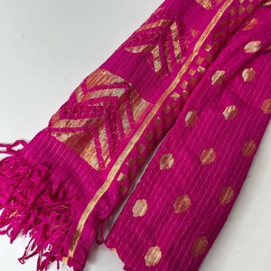 Puede incluir: Una bufanda de color rosa y dorado con flecos. La bufanda tiene un dise&ntilde;o geom&eacute;trico con detalles dorados.