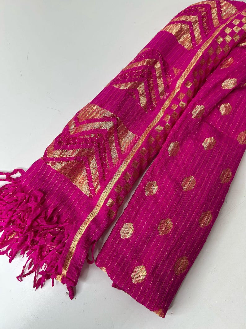 Puede incluir: Una bufanda de color rosa y dorado con flecos. La bufanda tiene un dise&ntilde;o geom&eacute;trico con detalles dorados.