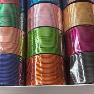 May include: A collection of colorful bangles in a variety of shades, including blue, pink, green, orange, and gold. The bangles are stacked neatly in a white tray, showcasing their shiny, metallic finish. The image highlights the diverse color palette and the reflective quality of the jewelry.