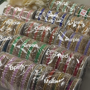 May include: A collection of cylindrical sets of bangles wrapped in clear plastic, each featuring a central colored band flanked by gold-toned, glitter-textured bangles. Labels identify colors including Peach, Pista, Blue, Haldi, Yellow, Orange, Black, Green, Purple, Red, Bpink, and Multi.