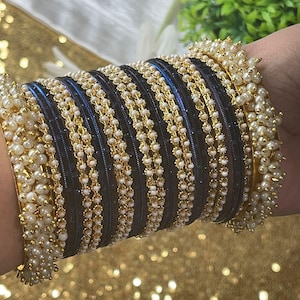 May include: A set of black and gold bangles with white pearl accents. The bangles are stacked on a person's wrist.