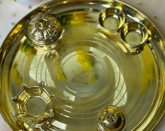 May include: A gold-colored metal puja thali with a circular design. The thali includes small bowls, a decorative container, and a small lamp. The background features a white pot with a green plant and yellow flowers.