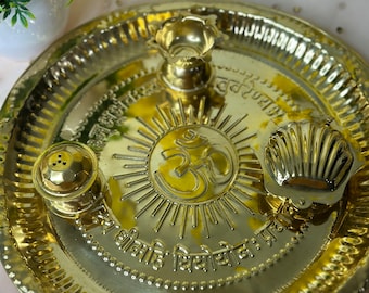 May include: A gold-colored metal puja thali with embossed religious symbols and text. The thali features a central sunburst design with the Om symbol, a small oil lamp, a shell-shaped container, and a small container with holes.