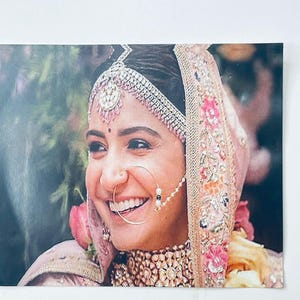 May include: A smiling person wearing traditional Indian wedding attire. The person is adorned with a jeweled headpiece, nose ring, and necklace. The outfit includes a pink and gold veil with floral embroidery.