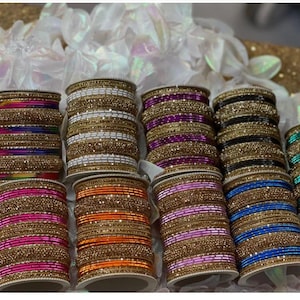 May include: A collection of colorful bangles in various stacks. The bangles feature gold-colored bands with embedded embellishments, and the stacks showcase a variety of colors, including pink, orange, blue, and green. The background is a mix of white and gold.