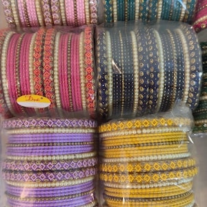 Puede incluir: Una colección de pulseras coloridas en varios tonos, incluyendo rosa, verde azulado, azul marino, morado y amarillo. Las pulseras están adornadas con patrones decorativos y perlas de imitación. La hora 3:15 PM es visible en la imagen.