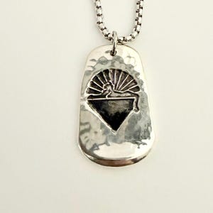 May include: A silver pendant necklace with a hammered texture. The pendant features a stylized design of a reclining figure beneath a sunburst and a diamond shape. The necklace chain is a box link style.