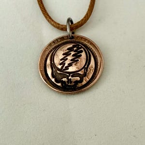 May include: A copper-colored pendant featuring a skull design with a lightning bolt. The pendant is attached to a brown cord necklace. The words "IN GOD WE TRUST" are visible on the coin.