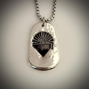 May include: A silver-toned dog tag pendant with a hammered texture, featuring a detailed black and silver emblem of a reclining figure with a sunburst design. The pendant hangs from a silver chain.
