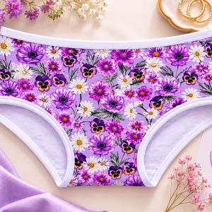 May include: A pair of purple underwear with a floral pattern featuring purple, white, and yellow flowers. The underwear has a white trim and is set against a light background with floral accents.