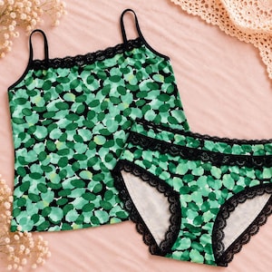 Puede incluir: Un conjunto de camiseta de tirantes y ropa interior a juego con un estampado abstracto de guijarros verdes y negros. Ambas piezas presentan un delicado borde de encaje negro en los tirantes y en las aberturas de las piernas.