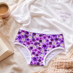 May include: A pair of purple underwear with a floral pattern of white and purple flowers. A white t-shirt with the text "Good things take time" and a heart is next to the underwear. A cup of coffee and a book are also in the image.