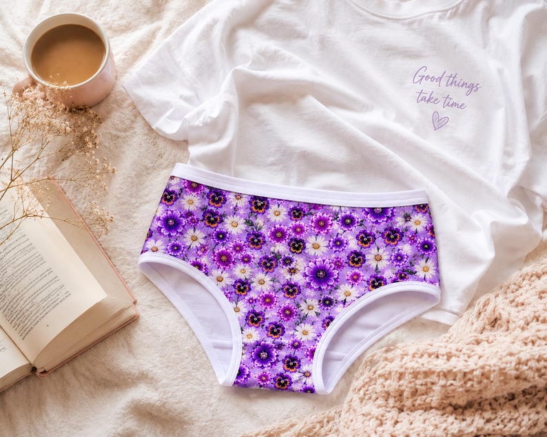 May include: A pair of purple underwear with a floral pattern of white and purple flowers. A white t-shirt with the text "Good things take time" and a heart is next to the underwear. A cup of coffee and a book are also in the image.