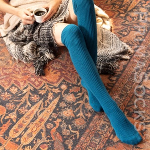 May include: A pair of teal blue thigh-high socks with a ribbed texture. The socks are worn by a person sitting on a patterned rug.