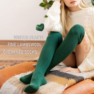 May include: Green over-the-knee socks made of fine lambswool. The socks have a ribbed texture and are shown on a model sitting on a sofa. "Winter Beauty" and "Fine Lambswool Overknee Socks" are written in white text on a brown background.