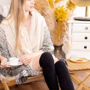 K&ouml;nnte beinhalten: Eine Frau in einem wei&szlig;en und schwarzen Strick&uuml;berwurf sitzt auf einem Holzstuhl mit einer Tasse Kaffee in der Hand. Sie tr&auml;gt schwarze Oberschenkelstr&uuml;mpfe und hat lange blonde Haare.
