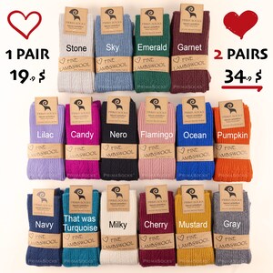 May include: A variety of 12 pairs of fine lambswool socks in different colors, including stone, sky, emerald, garnet, lilac, candy, nero, flamingo, ocean, pumpkin, navy, turquoise, milky, cherry, mustard, and gray. The socks are arranged in two rows of six. Each pair is labeled with the color name and the words "Fine Lambswool".