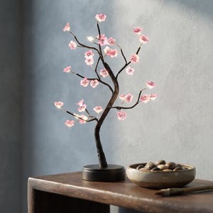 May include: A decorative LED lamp shaped like a bonsai tree with a dark brown trunk and branches adorned with small, glowing pink cherry blossom flowers. The lamp features a circular black base and emits a soft light from the flower centers.