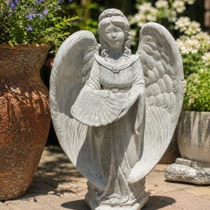 May include: A gray stone angel statue featuring large, detailed wings, a flowing robe with a small circular brooch at the neckline, and a fan held in both hands. The figure stands on a hexagonal base with a textured, weathered finish throughout.