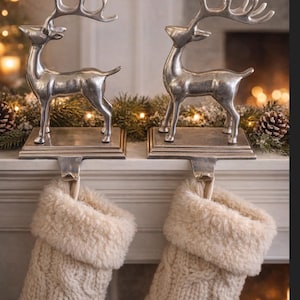 Puede incluir: Dos colgadores de calcetines de reno plateados con calcetines de punto color crema debajo. Los renos están sobre una repisa decorada con vegetación y luces, creando una escena festiva navideña.