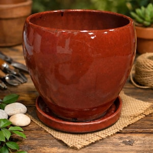 May include: A glossy, red-brown ceramic plant pot with a matching saucer. The pot has a rounded shape and sits on a burlap square. Other gardening tools and supplies are visible in the background. The pot is designed for indoor or outdoor use.