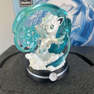 May include: A white and silver figurine of a cartoon fox-like creature, surrounded by translucent blue ice and a snowflake. The figurine sits on a black base with a silver trim and a logo. The background features a blue illustration and the "EMG Studios" logo.