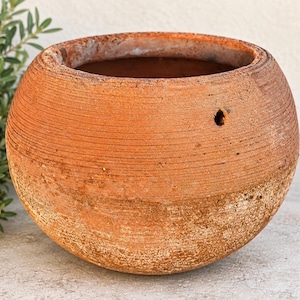 May include: A round terracotta planter with a textured, ribbed surface and a weathered, earthy orange finish. The pot features a wide, open top and a single small circular hole on the upper side, showcasing a rustic, porous clay construction.