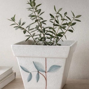 Könnte beinhalten: Ein quadratischer, weißer Keramik-Blumentopf mit Craquelé-Finish, verziert mit einem gemalten Design aus blauen Blättern und einem braunen Stiel. Der Topf enthält eine grüne Blattpflanze. Der Topf steht auf einer Holzoberfläche.