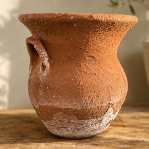 May include: A rustic, terracotta-colored vase with a wide, flared rim and two small handles. The textured surface has a weathered appearance with white mineral deposits near the base. The vase sits on a wooden surface, suggesting a vintage or antique aesthetic.