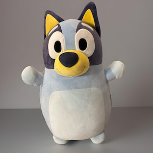 May include: A plush toy shaped like a blue dog with large white oval eyes, a yellow muzzle, and a black nose. The character has dark blue ears with yellow inner sections, a light blue body, and a pale blue belly patch with outstretched arms.