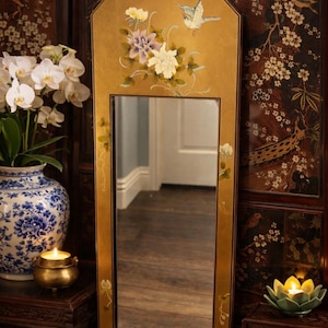 以下が含まれることがあります： 花と鳥のアートワークが施された長方形の金色のフレームの鏡。鏡は、花の模様が施された暗い木製のスクリーンの前に配置されています。白い花とろうそくが入った青と白の花瓶が前景にあります。