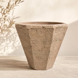 May include: A geometric, light brown planter with a textured surface. The planter has a wide, open top and a faceted, tapered design. The neutral color and modern shape make it suitable for various plants and decor styles.