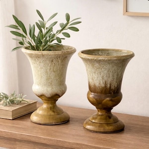 May include: Two decorative urn-shaped vases with a weathered, aged appearance. One vase holds green leafy branches. The vases have a two-tone color scheme, with a light beige upper portion and a darker brown base. They sit on a wooden surface.