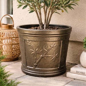 May include: A large, bronze-colored outdoor planter with a decorative bamboo branch design. The planter holds a small olive tree with green leaves. A woven lantern and a stack of books are in the background.