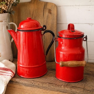 May include: A red enamel coffee pot and milk jug set. The coffee pot has a black handle and spout, while the milk jug features a wooden handle. Both items are on a wooden surface.