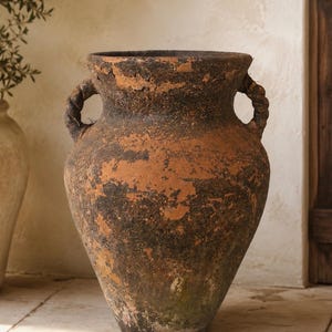 May include: A large, weathered terracotta vessel with a wide mouth and two twisted rope-style handles. The exterior features a mottled, textured finish with patches of dark brown, black, and earthy orange tones, showing signs of age and wear.