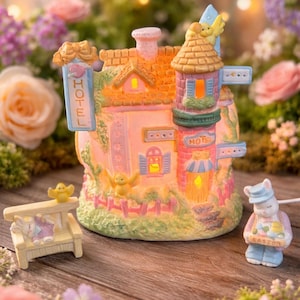 May include: A pastel Easter-themed ceramic house with a "HOTEL" sign, a small bench, and a bunny figurine holding a sign that says "EASTER". The house has a pink facade, orange roof, and blue accents. The scene is set on a wooden surface with flowers.