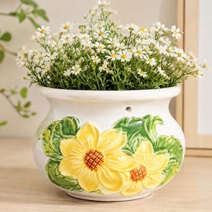 Peut inclure: Un pot de fleurs en céramique blanche avec des motifs de tournesols jaunes et des feuilles vertes. Le pot est rempli de petites fleurs blanches à centres jaunes. Le pot est sur une surface en bois.