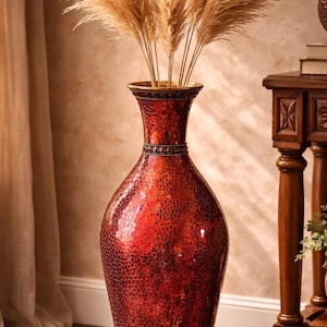 May include: A tall, red decorative vase with a textured mosaic pattern. The vase is filled with dried, tan-colored ornamental grass. The vase has a gold rim and sits on a wooden floor. A wooden table is in the background.