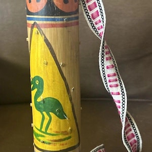 May include: A cylindrical bamboo rattle with hand-painted designs. The rattle features a green bird on a yellow background, orange circles, and colorful stripes. A patterned strap is attached to the top.